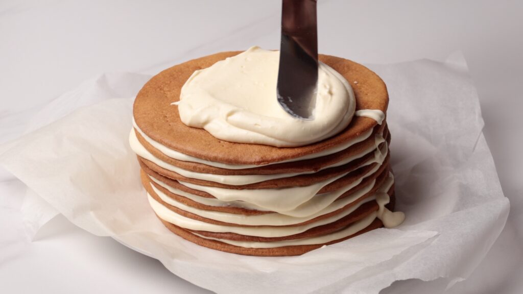 A spatula spreads creamy white frosting on top of a stacked layered cake made of thin, round cookies, set on crumpled parchment paper against a white background.