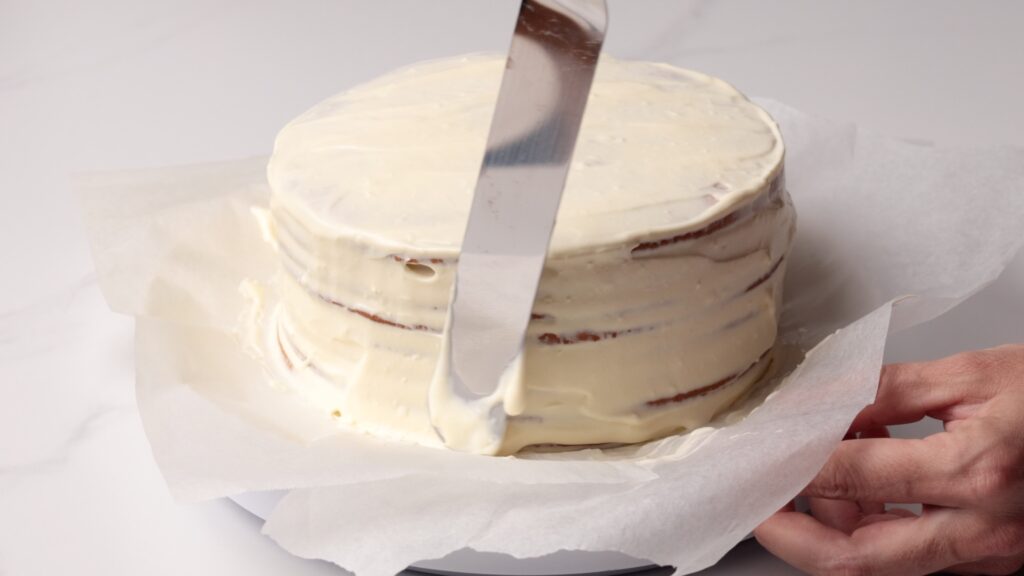 A hand uses an offset spatula to spread white frosting on a layered cake placed on parchment paper over a plate.