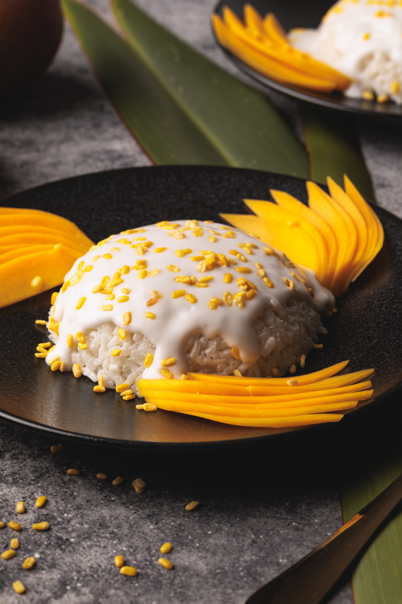A plate of Thai mango sticky rice topped with coconut cream and sprinkled with yellow mung beans, garnished with sliced mango, served on a black plate with green leaves beneath.