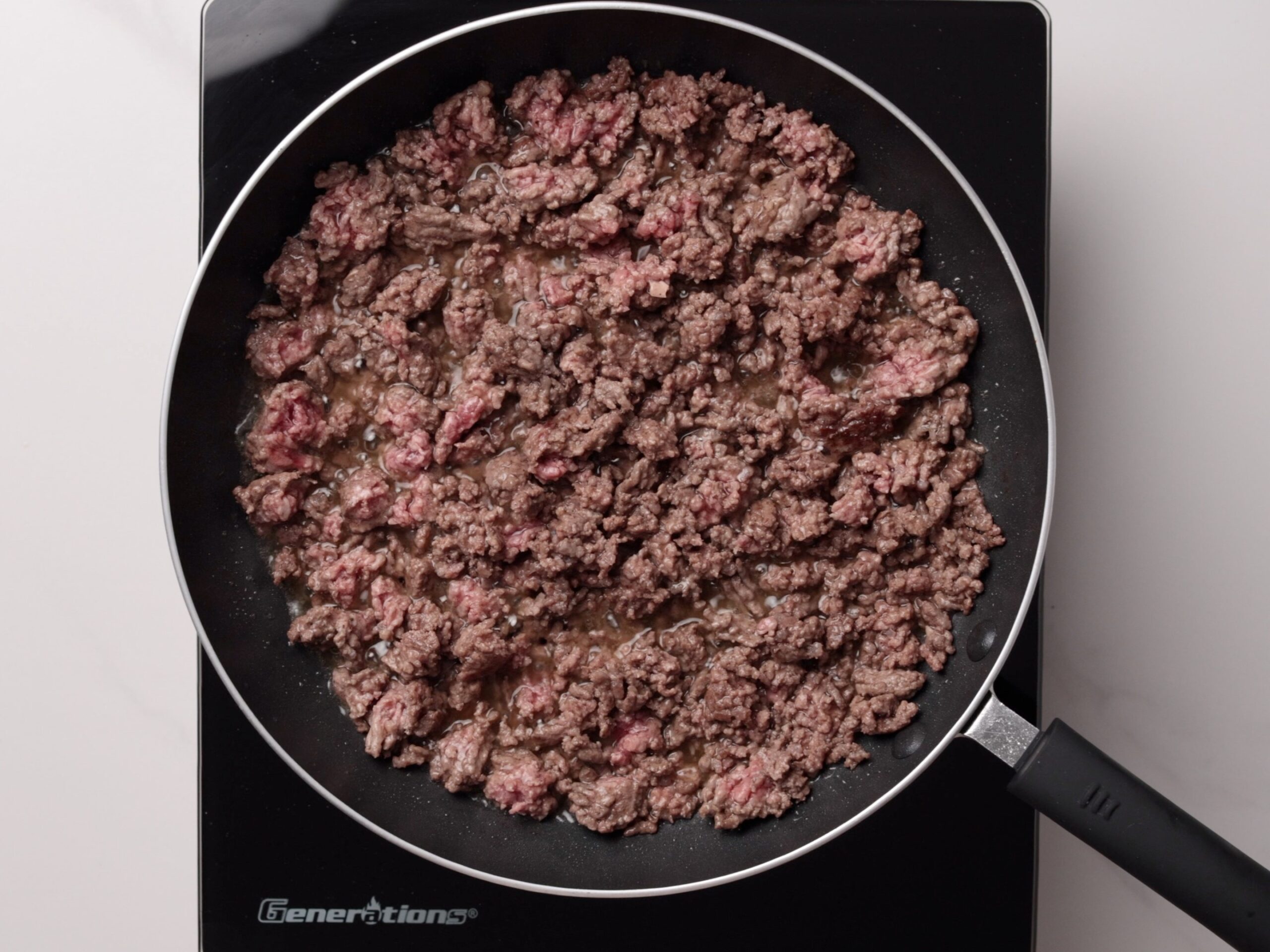 Ground beef is being browned in a frying pan on a stovetop. Some pieces are still pink while others are cooked brown, and there is visible liquid in the pan. The stovetop is black with a handle visible.