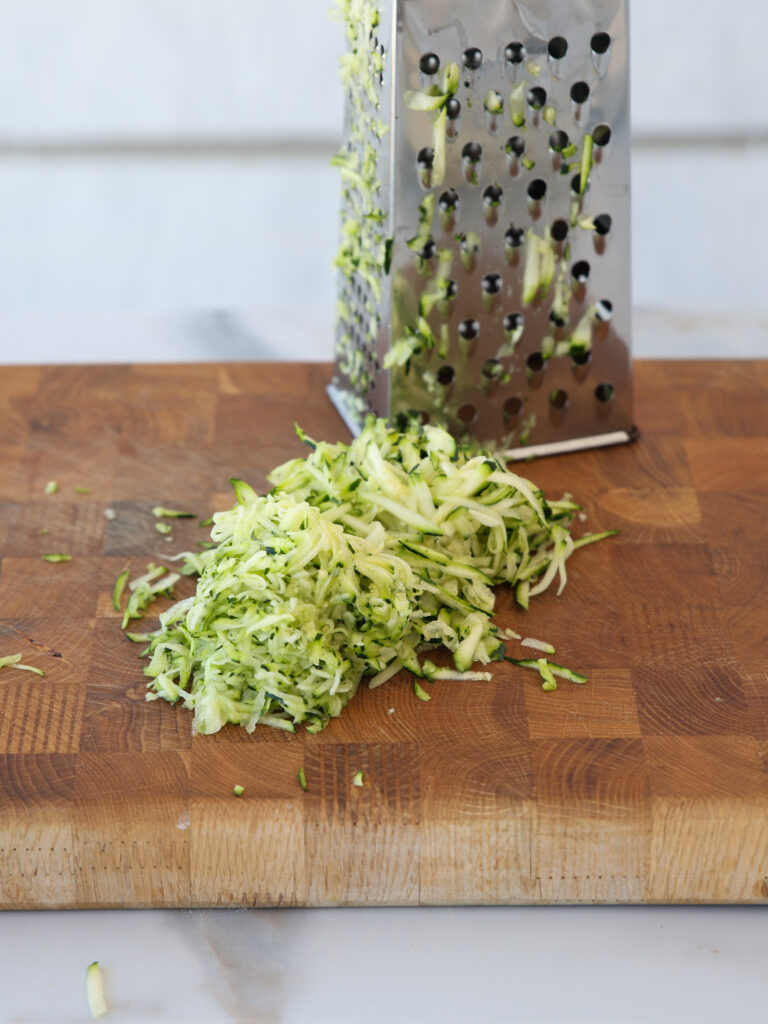 A pile of freshly grated zucchini sits on a wooden cutting board next to a metal box grater.