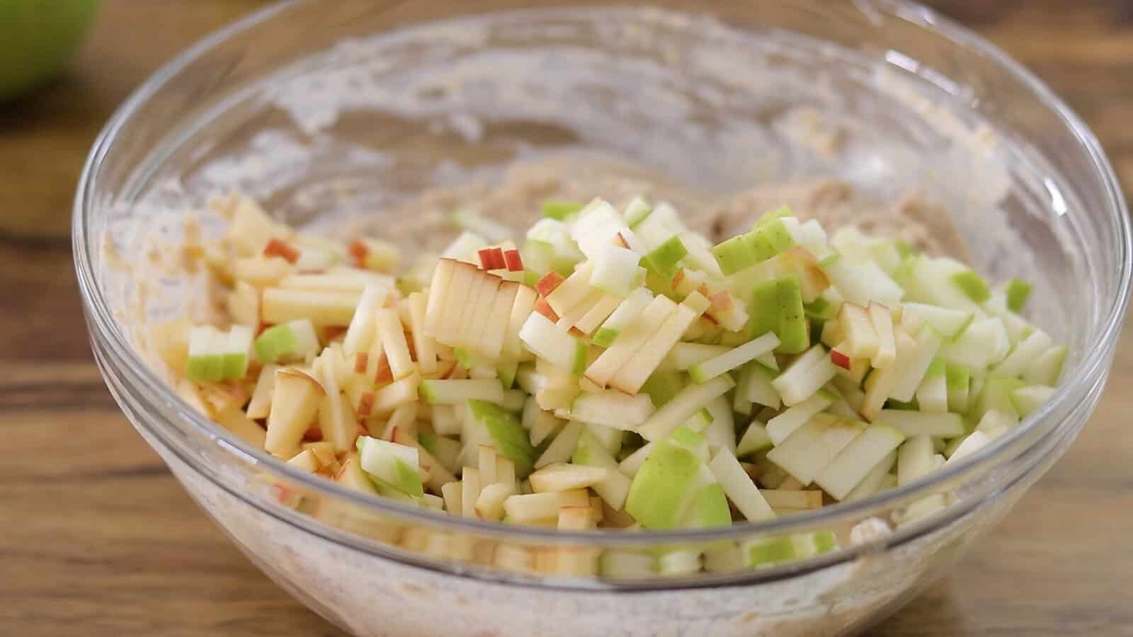 A clear glass bowl filled with chopped green and red apples mixed into a light-colored batter, sitting on a wooden surface.