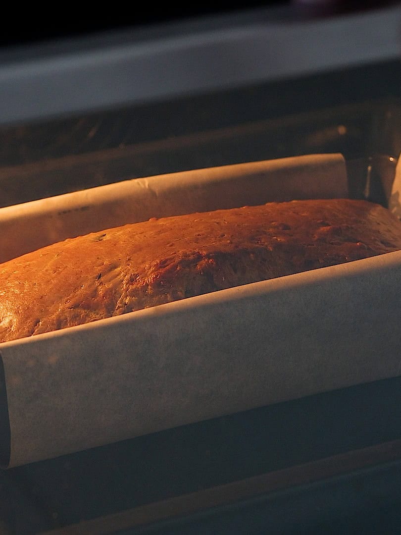 A loaf of bread or cake is baking in an oven, inside a rectangular pan lined with parchment paper. The top is golden brown and rising as it cooks.