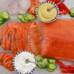 Slices of gravlax salmon arranged on a cutting board with sliced cucumbers, tomatoes, red onions, red bell peppers, and bowls of mustard and sour cream with spoons.
