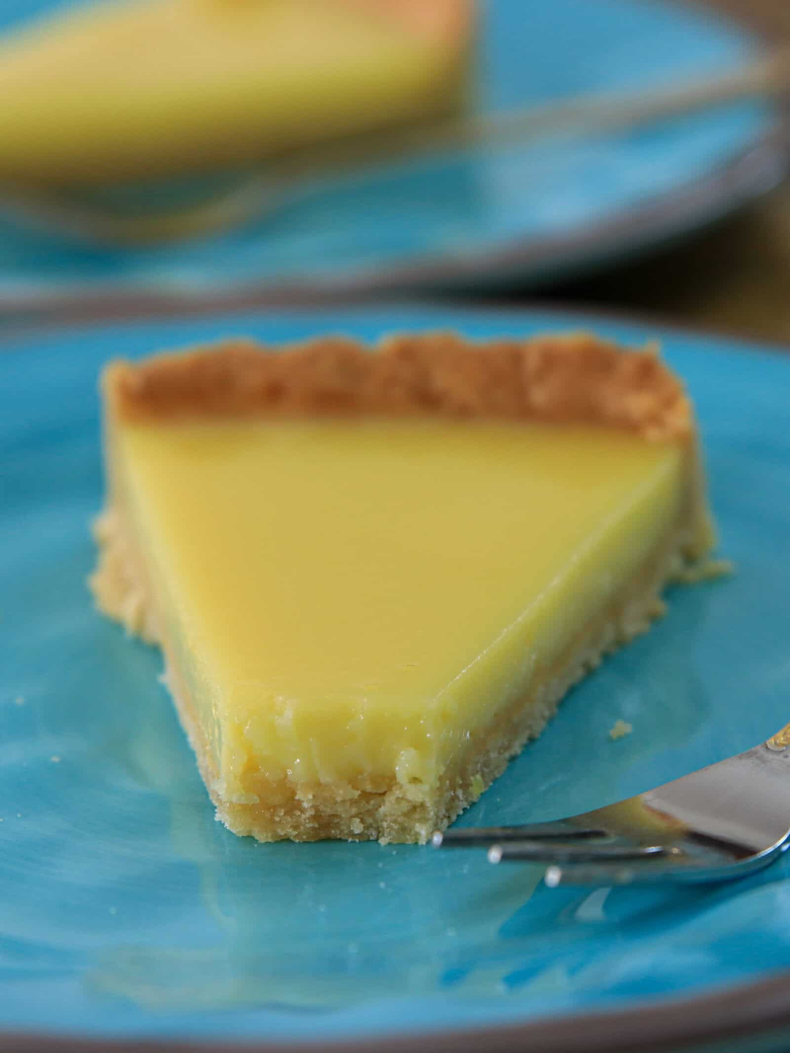 A close-up of a slice of lemon tart with a golden crust, served on a blue plate with a fork beside it. Another slice is blurred in the background on a similar plate.