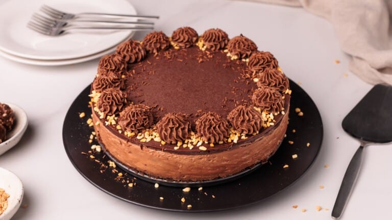 A Nutella cheesecake (no-bake) with chocolate frosting and piped rosettes, topped with chocolate sprinkles and chopped nuts around the edge, sits on a black plate. Plates, forks, and a cake server are nearby on a white surface.