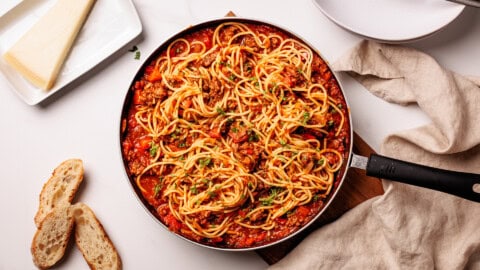 A skillet filled with spaghetti in tomato-meat sauce sits on a table, surrounded by slices of bread, a wedge of parmesan cheese on a plate, and a beige linen napkin.