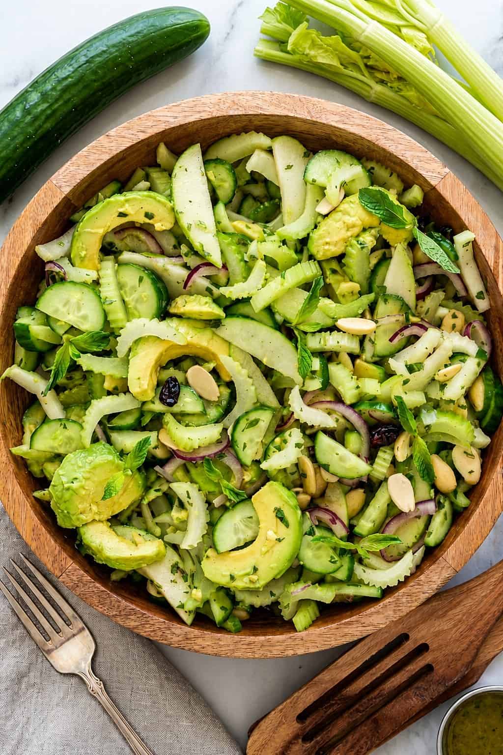 A wooden bowl filled with fresh green celery and cucumber salad featuring sliced avocado, cucumber, celery, fennel, red onion, nuts, and fresh herbs. Whole cucumber and celery stalks are nearby, along with salad utensils and a fork.