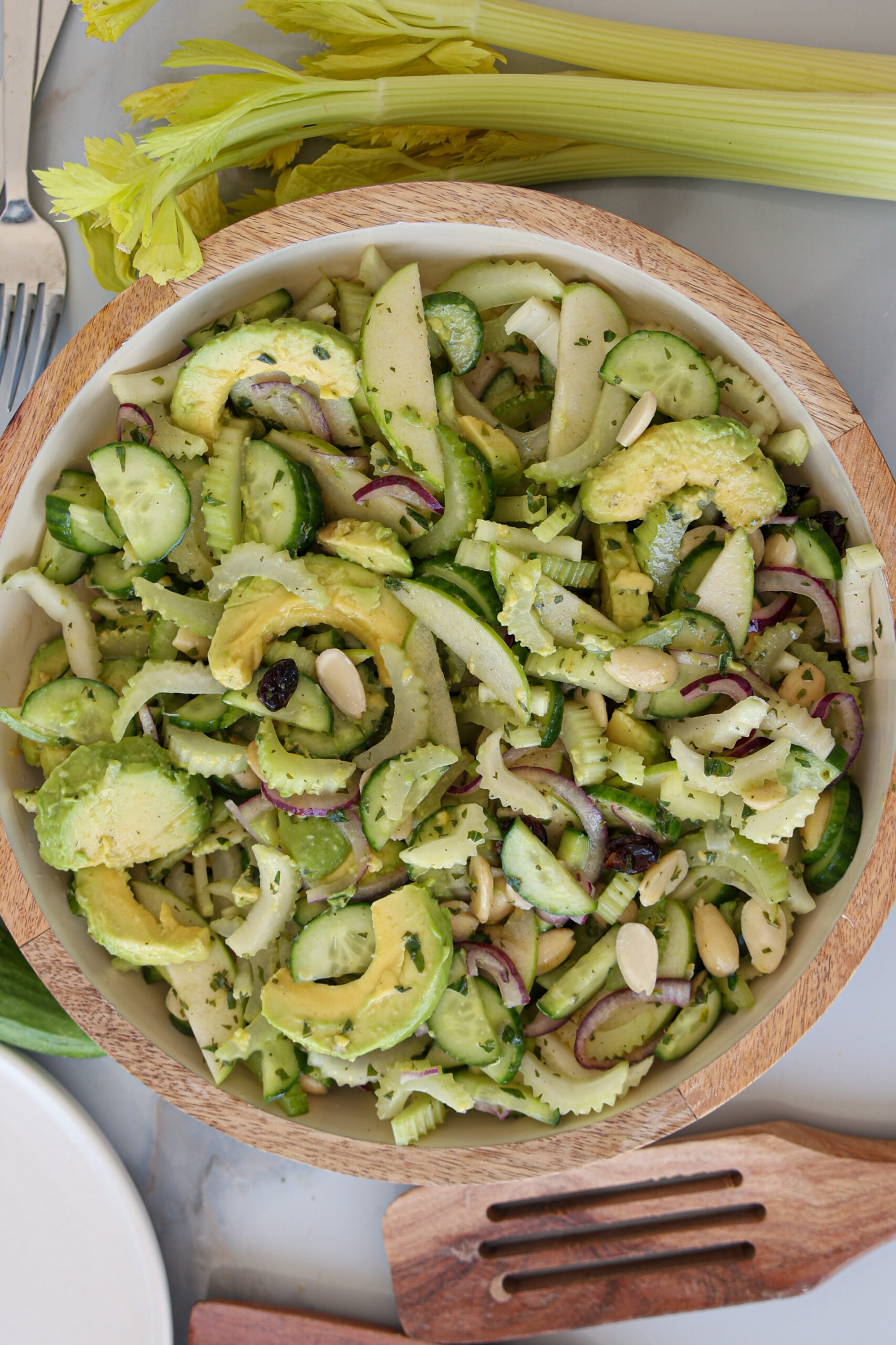 A wooden bowl filled with a fresh celery and cucumber salad containing sliced cucumber, avocado, red onion, celery, and slivered almonds, placed on a table with celery stalks and utensils nearby.