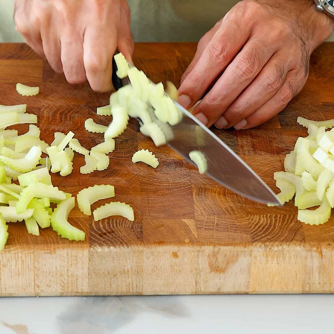 A person’s hands are slicing celery with a large knife on a wooden cutting board. Chopped celery pieces are scattered on the board.