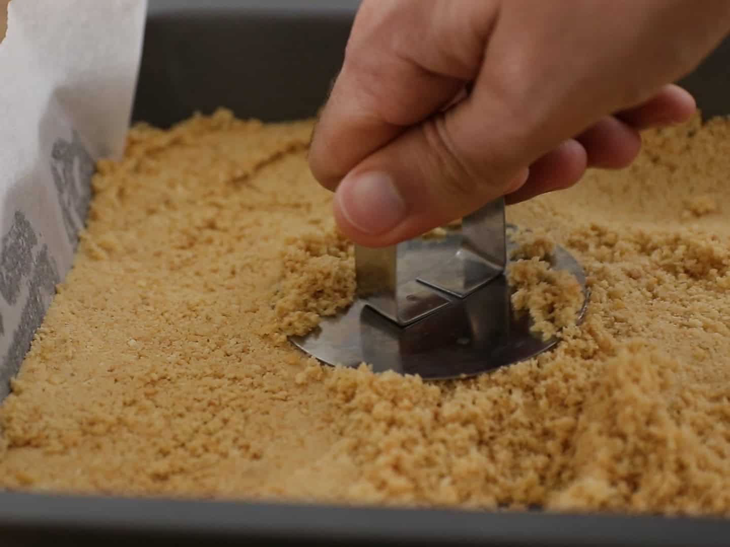 A hand presses a metal tool onto a crumbly biscuit base in a parchment-lined baking pan, flattening the mixture evenly.