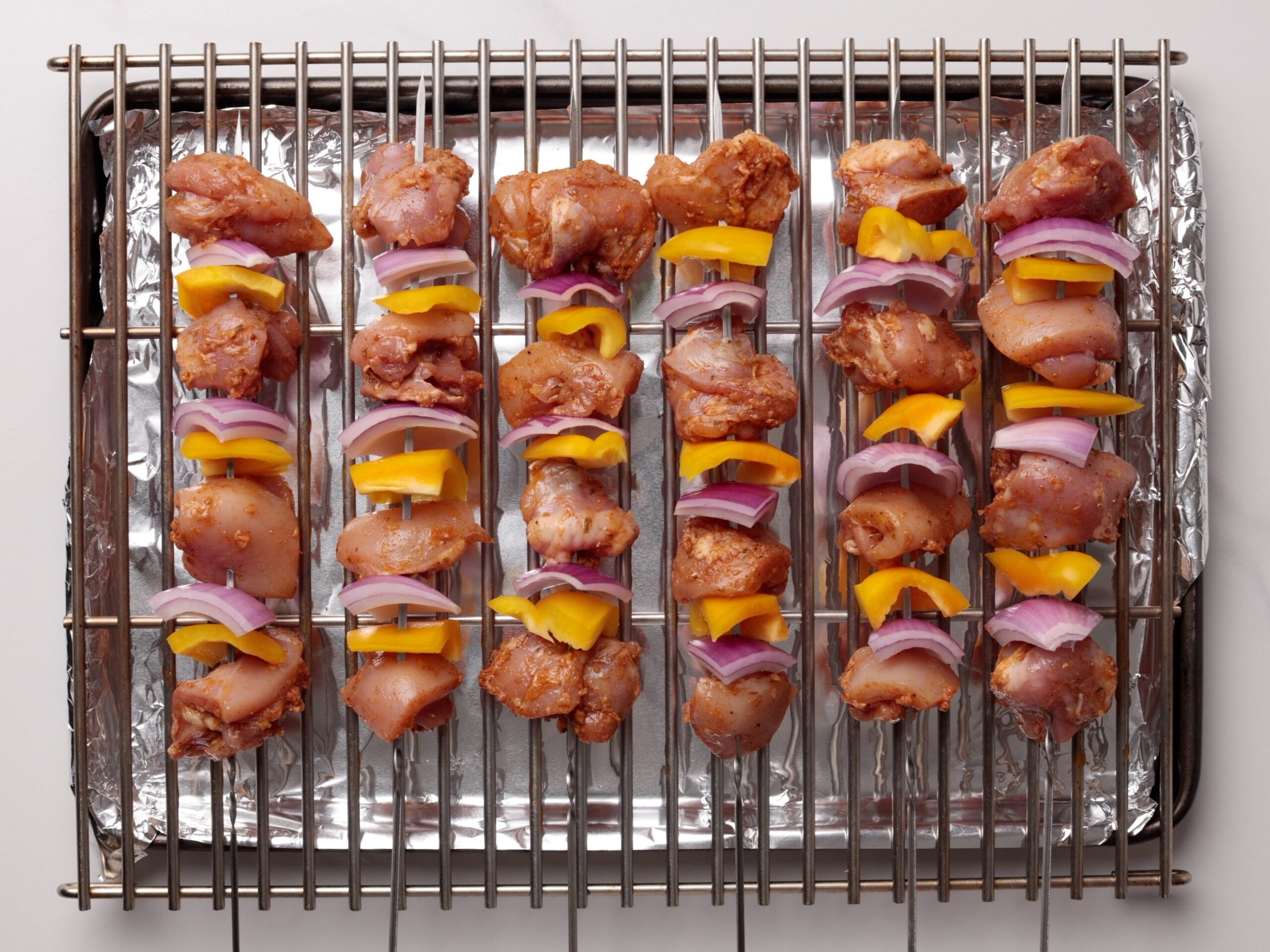 Raw chicken, yellow bell pepper, and red onion pieces are skewered on metal sticks and arranged on a grill rack over a foil-lined baking tray, ready to be cooked.