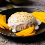 A dessert of mango sticky rice topped with coconut cream and sprinkled with yellow mung beans, served with sliced ripe mango on a black plate.