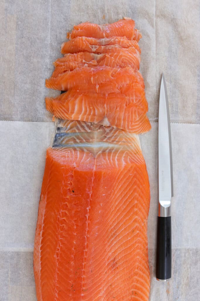 A fillet of cured salmon is partially sliced on parchment paper with a sharp knife placed alongside it. The vibrant orange-pink salmon contrasts with the neutral paper background.