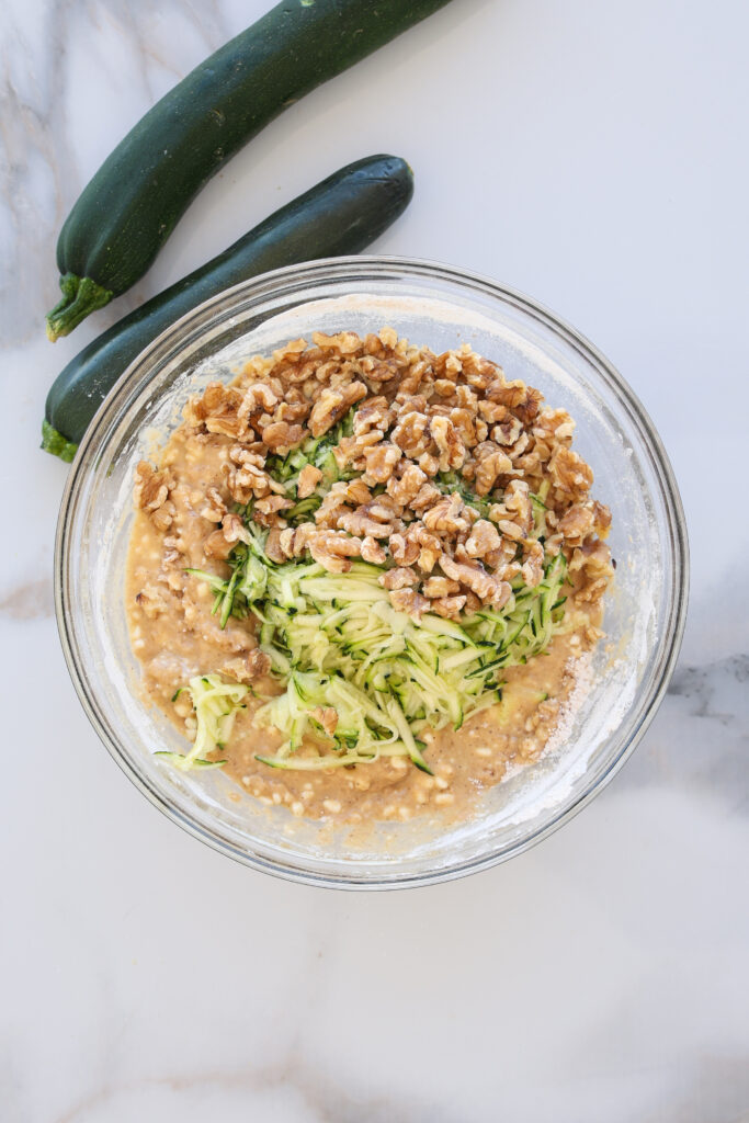 A glass bowl filled with zucchini bread batter, topped with shredded zucchini and chopped walnuts, sits on a marble surface. Two whole zucchinis are placed next to the bowl.