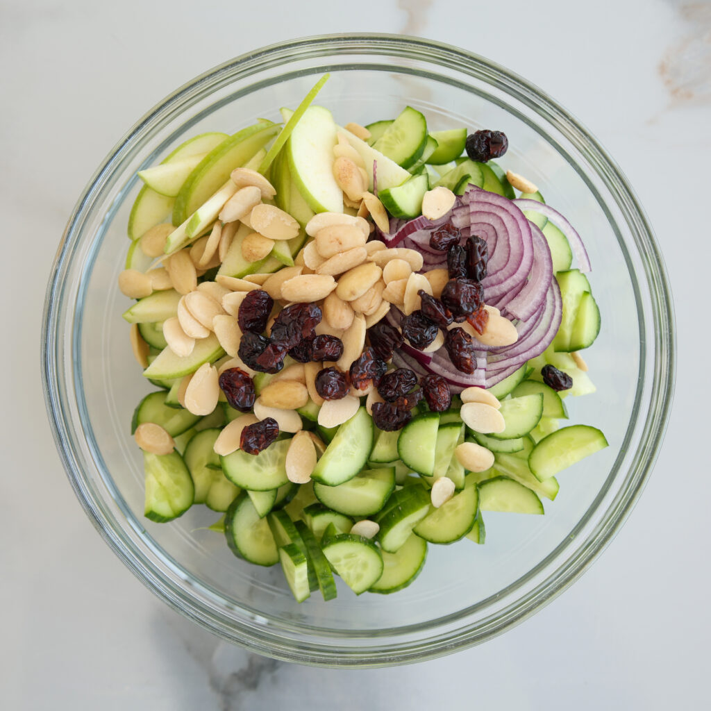 A glass bowl filled with sliced cucumbers, red onions, green apple slices, topped with almonds and dried cranberries, viewed from above on a light surface.