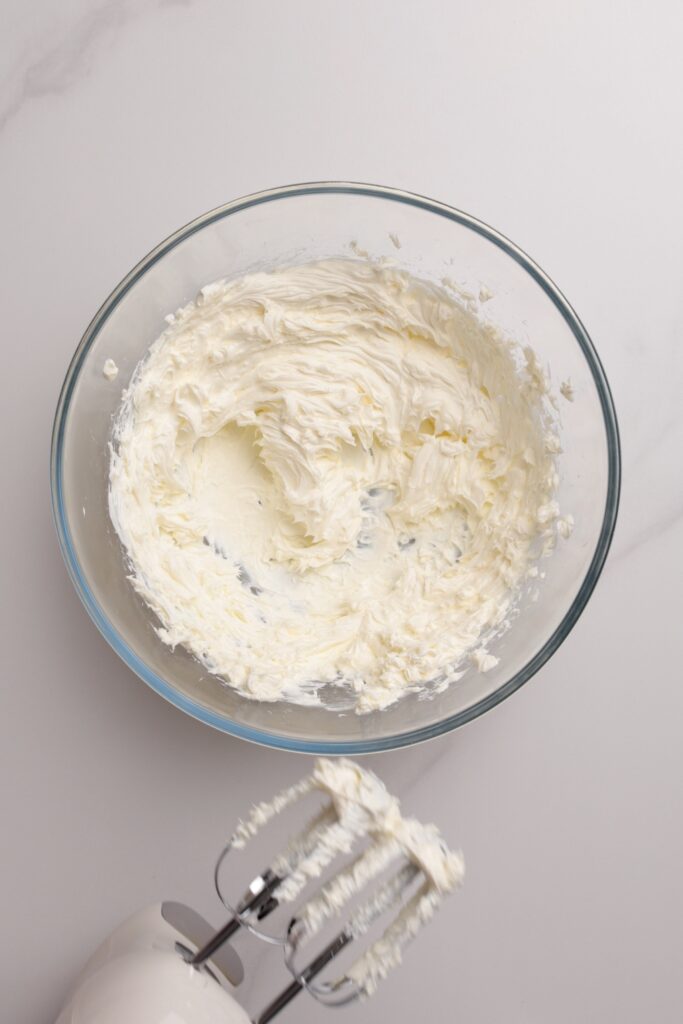 A hand mixer with creamy frosting on its beaters is shown next to a glass bowl of frosting on a marble surface.