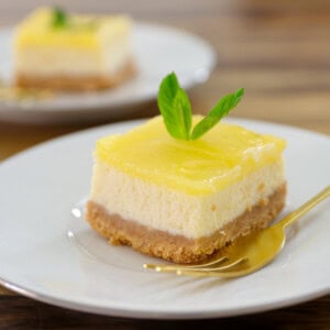 A square slice of lemon cheesecake (lemon cheesecake bars) with a crumbly base, creamy layer, and yellow topping, garnished with a mint leaf, sits on a white plate with a gold fork. Another slice is blurred in the background.