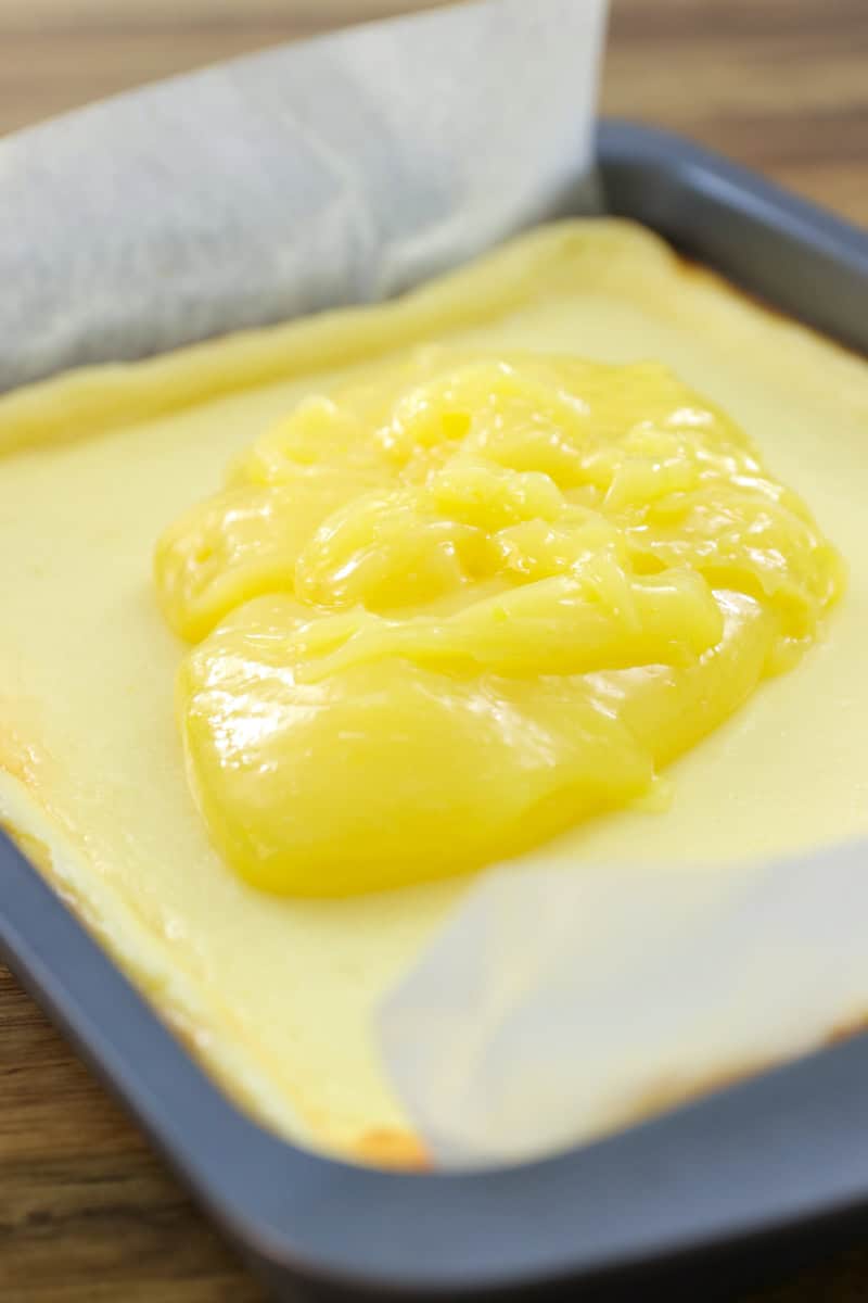 A baking pan lined with parchment paper holds a baked square cheesecake topped with a large dollop of bright yellow lemon curd.