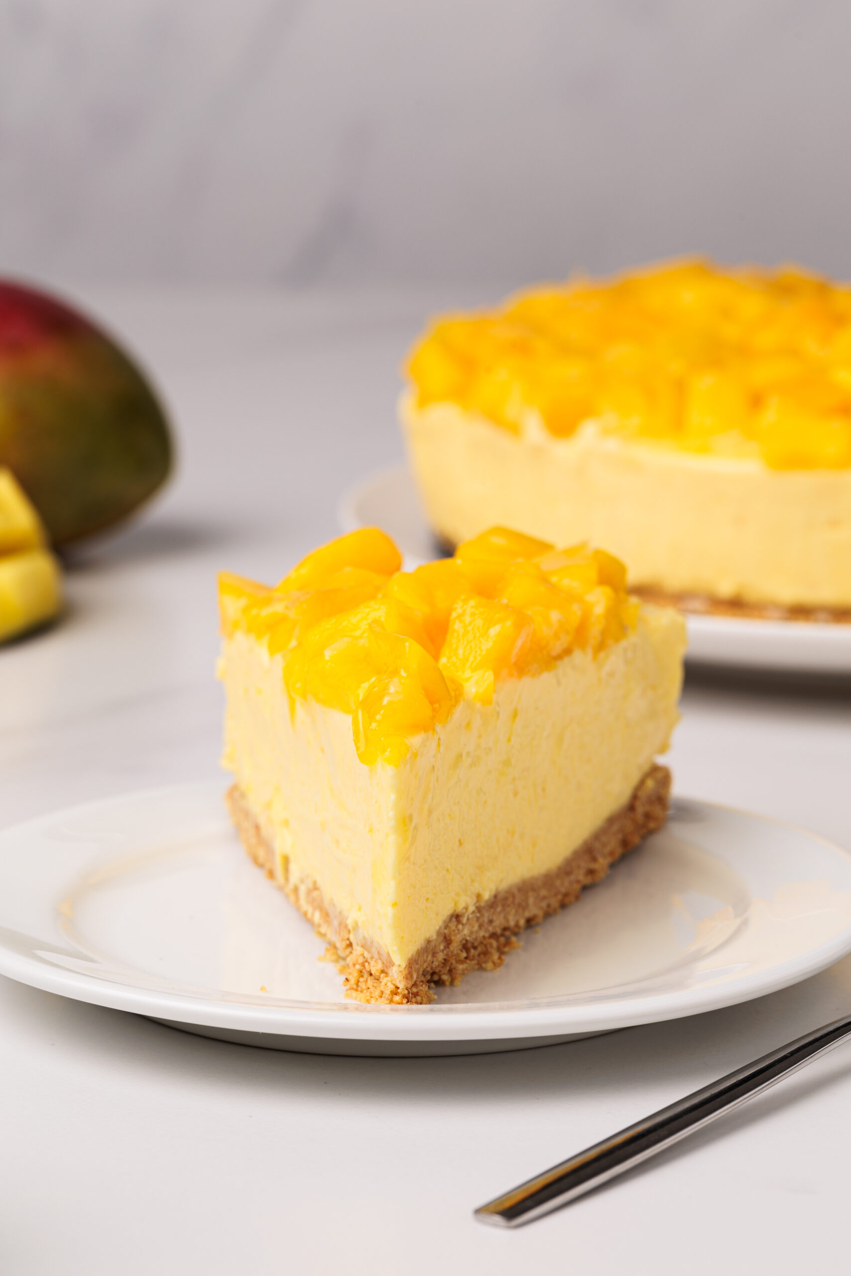 A slice of creamy mango cheesecake sits on a white plate, topped with fresh mango chunks. A whole mango is blurred in the background.