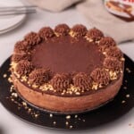 A chocolate cake with chocolate frosting, decorated with piped swirls and chopped nuts around the edge, sits on a black plate. Plates and a Nutella jar are visible in the background.