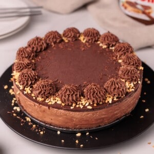 A chocolate cake with chocolate frosting, decorated with piped swirls and chopped nuts around the edge, sits on a black plate. Plates and a Nutella jar are visible in the background.
