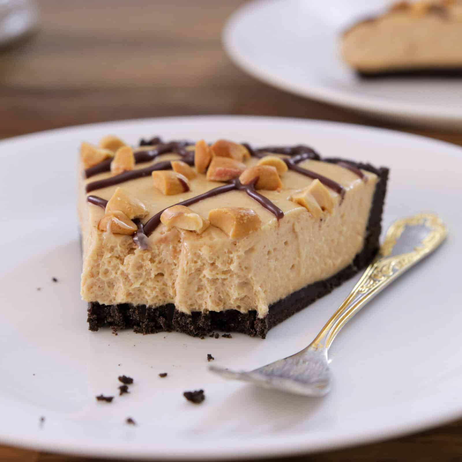 A slice of creamy no-bake peanut butter pie with a chocolate crust, topped with chopped peanuts and chocolate drizzle, sits on a white plate with a silver fork.