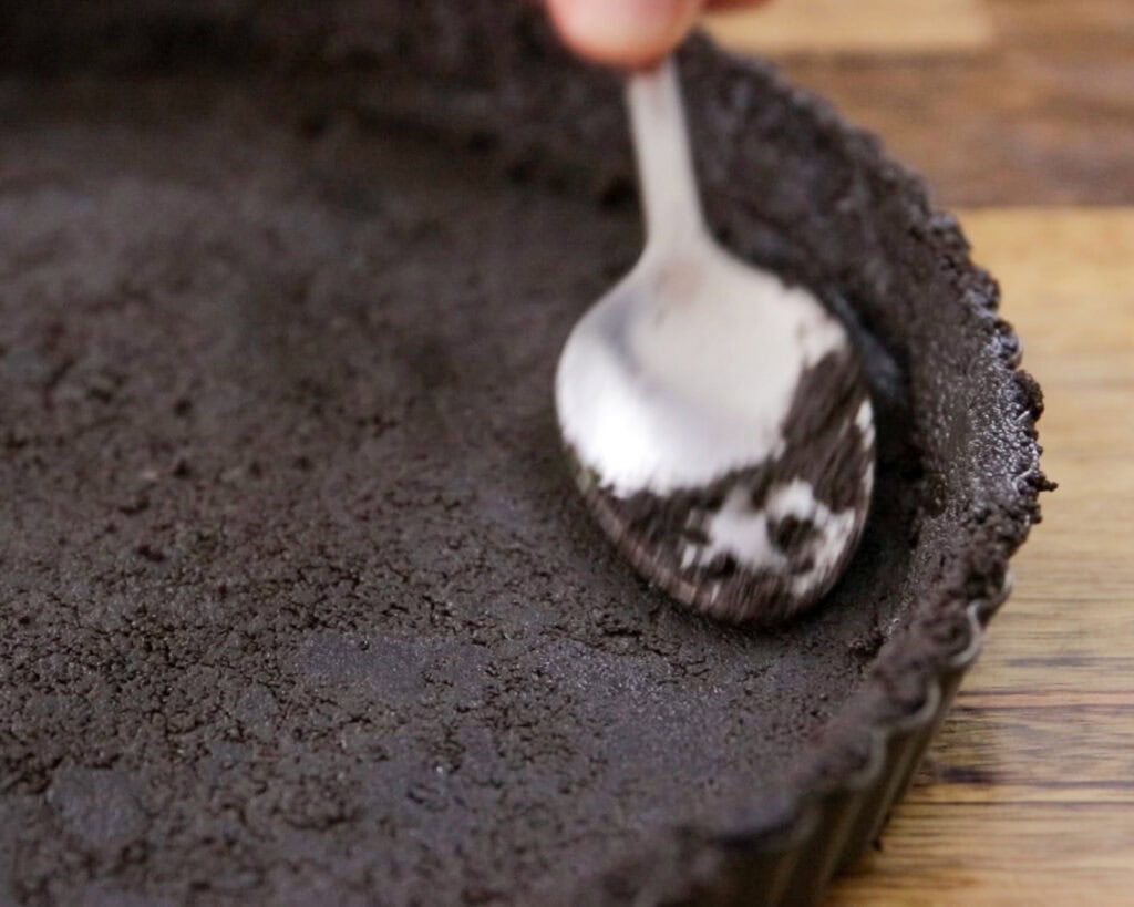 A hand uses a spoon to press down and smooth a dark chocolate cookie crust in a round tart pan on a wooden surface.