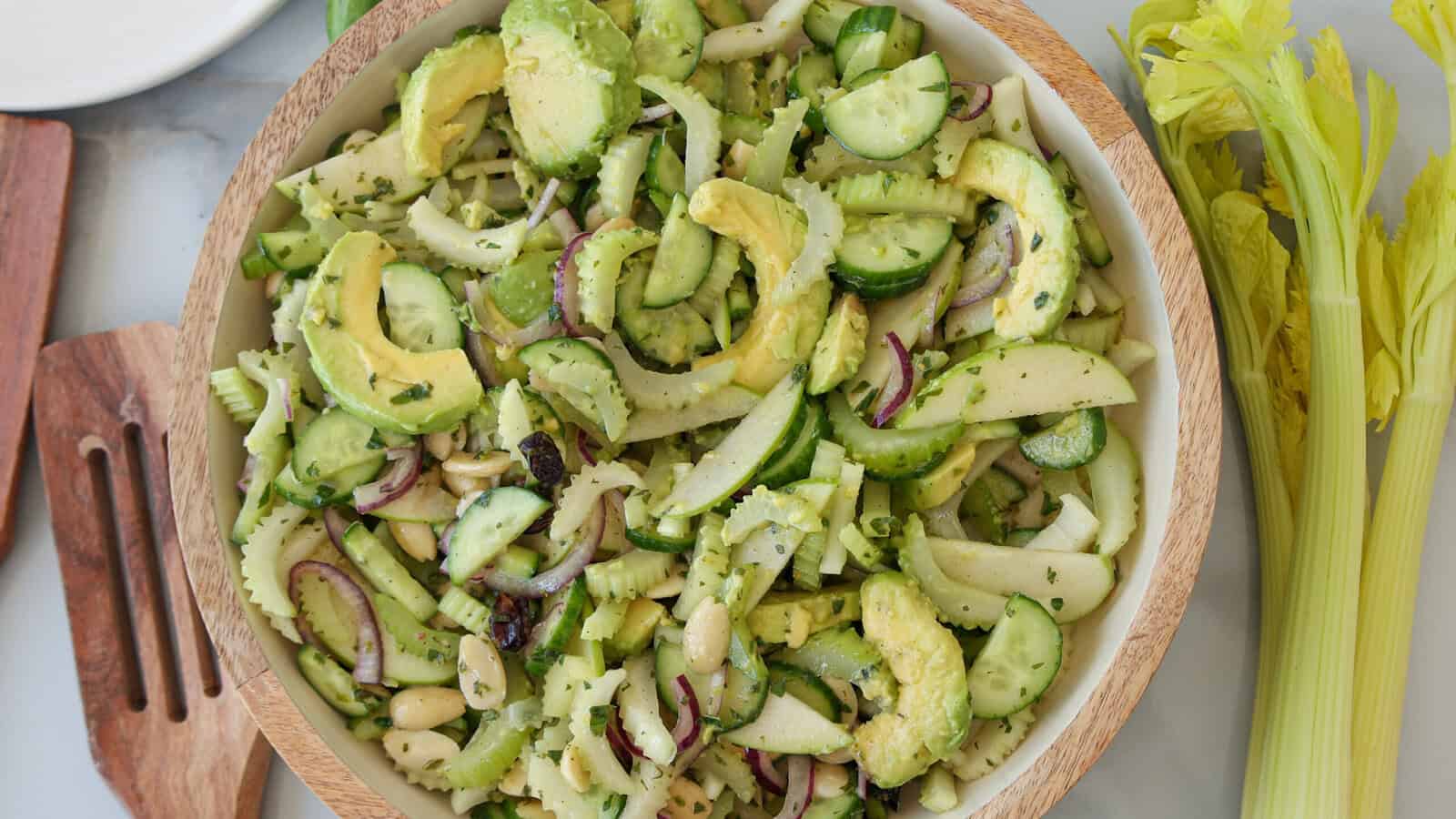 A wooden bowl filled with a fresh cucumber and celery salad containing sliced avocado, cucumber, fennel, red onion, celery, apple, and white beans, next to celery stalks and wooden utensils.