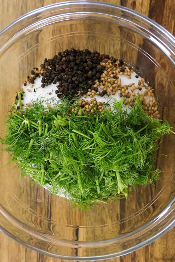 A glass bowl containing fresh dill, whole black peppercorns, coriander seeds, and a mound of white salt on a wooden surface.