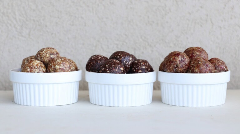 Three white ramekins, each filled with a stack of round energy date balls in different shades of brown, sit side by side on a white surface against a light textured background.