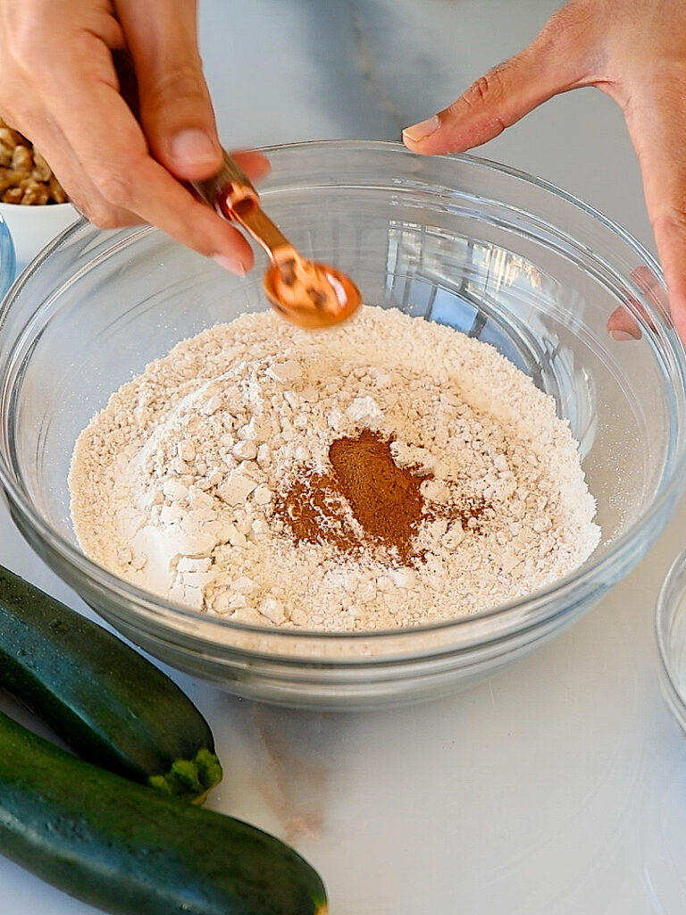A person adds ground cinnamon from a measuring spoon to a large glass bowl filled with flour. Two zucchinis and other small bowls are on the white countertop nearby.