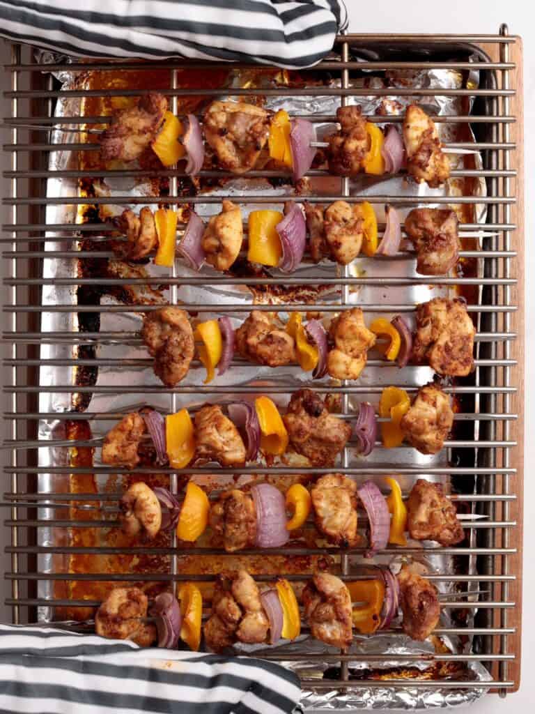 Five skewers with baked chicken Kabobs, yellow bell peppers, and red onions rest on a metal rack over a foil-lined baking tray. A person wearing striped oven mitts is holding the tray.