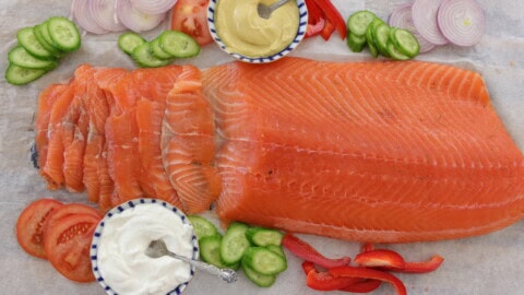 Sliced cured salmon (gravlax) fillet with bowls of mustard sauce and cream, surrounded by sliced cucumbers, red onions, tomatoes, and red bell peppers on a white surface.