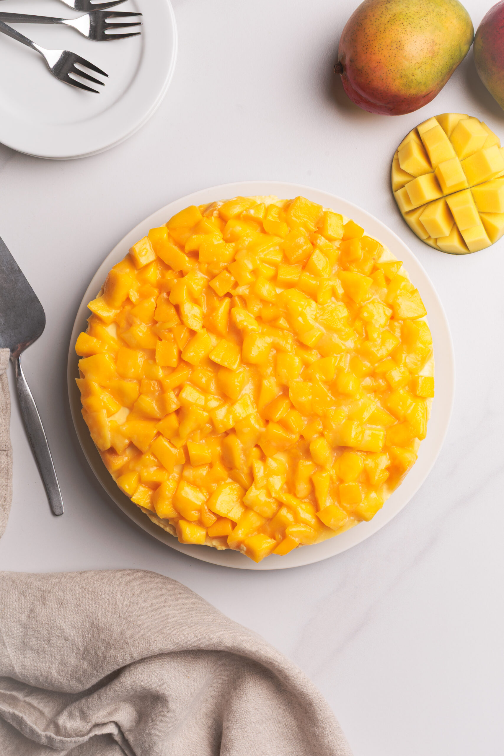 A delicious mango cheesecake topped with diced fresh mangoes sits on a white plate, with two whole mangoes and a slice of mango nearby.