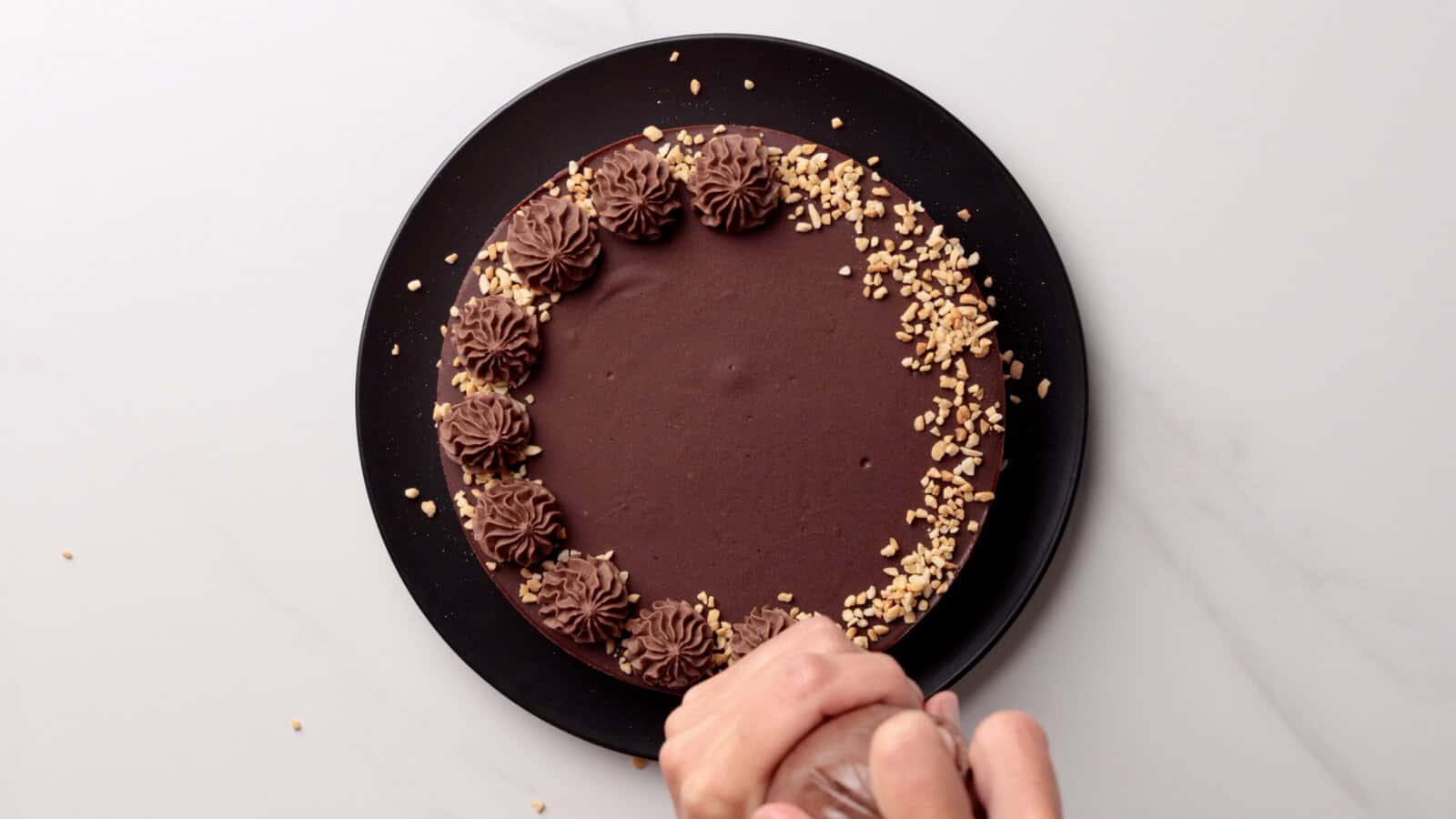 A hand decorates a no-bake nutella cheesecake with chocolate frosting swirls and chopped nuts on a black plate, viewed from above on a light surface.