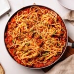 A skillet filled with spaghetti mixed in a rich tomato and meat sauce, garnished with chopped herbs, sits on a white table beside a napkin and empty plate.