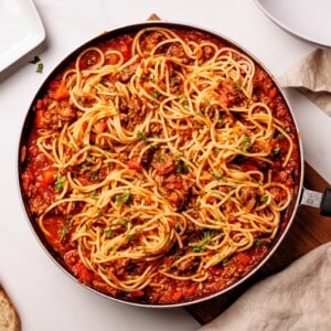 A skillet filled with spaghetti mixed in a rich tomato and meat sauce, garnished with chopped herbs, sits on a white table beside a napkin and empty plate.