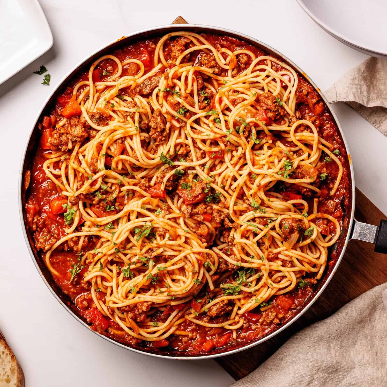 A skillet filled with spaghetti mixed in a rich tomato and meat sauce, garnished with chopped herbs, sits on a white table beside a napkin and empty plate.
