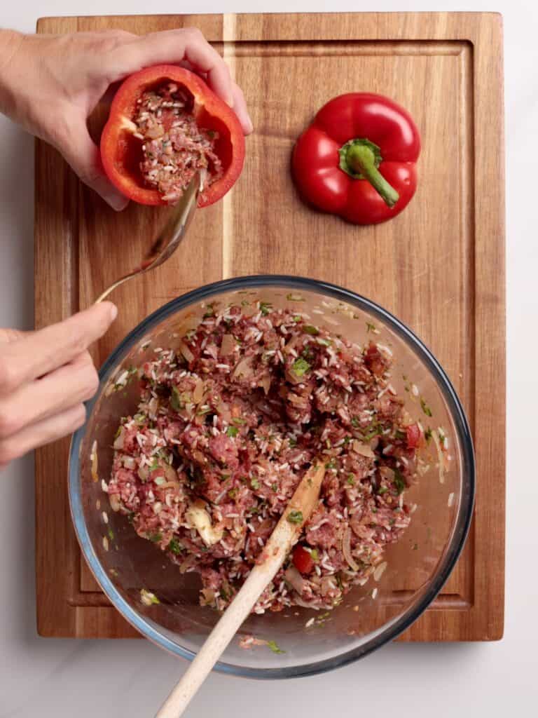 A person fills a red bell pepper with a mixture of ground meat, rice, and herbs from a glass bowl on a wooden cutting board; another whole bell pepper sits nearby.
