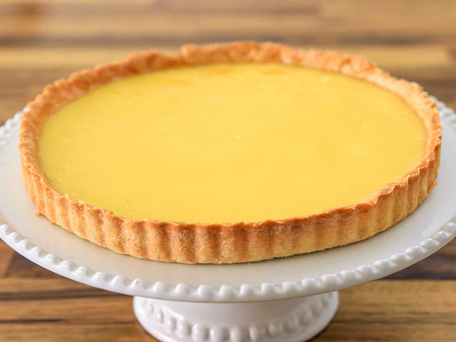 A French lemon tart with a golden, smooth filling and a crisp, fluted pastry crust sits on a white, decorative cake stand against a wooden background.