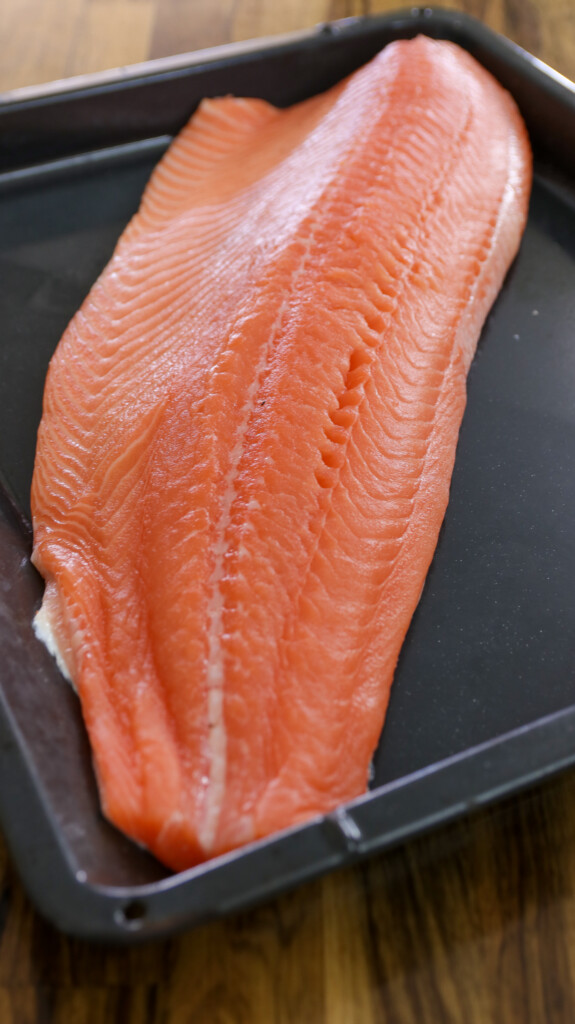 A large, raw salmon fillet with a bright orange-pink color lies on a black baking tray, placed on a wooden surface.