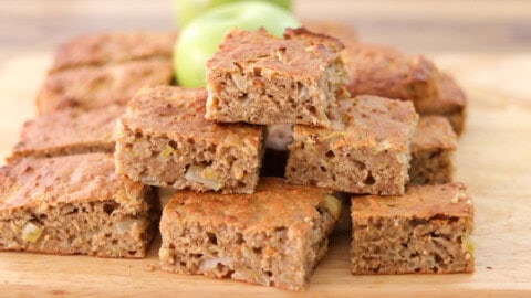 A stack of moist, square slices of homemade apple cake on a wooden board, with green apples blurred in the background. The cake appears dense with small pieces of apple visible inside.