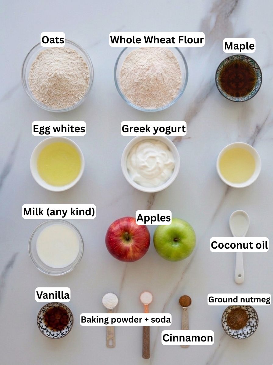 Top-down view of labeled ingredients for a recipe, including oats, whole wheat flour, maple syrup, egg whites, Greek yogurt, milk, apples, coconut oil, vanilla, baking powder and soda, cinnamon, and ground nutmeg.