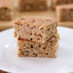 Two pieces of moist, homemade healthy apple cake are stacked on a white plate. The cake has a golden-brown crust and visible apple chunks inside. More cake pieces are blurred in the background.