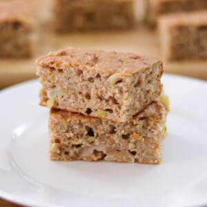 Two pieces of moist, homemade healthy apple cake are stacked on a white plate. The cake has a golden-brown crust and visible apple chunks inside. More cake pieces are blurred in the background.