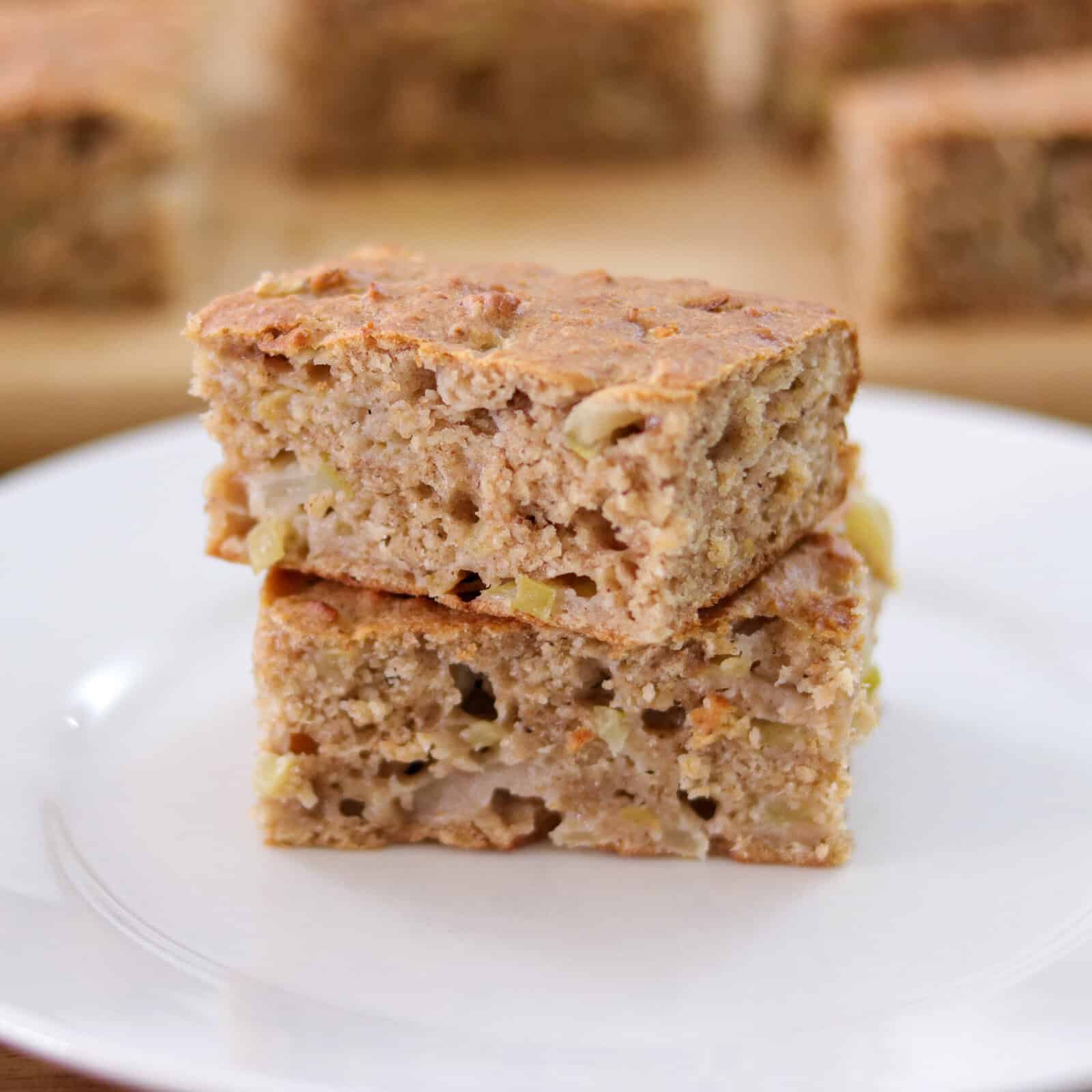 Two pieces of moist, homemade healthy apple cake are stacked on a white plate. The cake has a golden-brown crust and visible apple chunks inside. More cake pieces are blurred in the background.