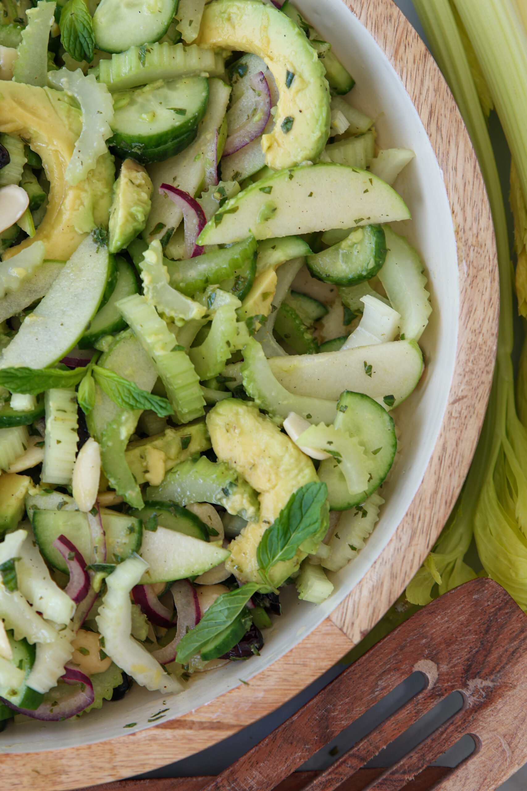A close-up of a fresh summer salad in a wooden bowl, featuring sliced avocado, cucumber, celery, green apple, red onion, and herbs, with a wooden serving utensil nearby.