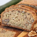 Sliced cottage cheese zucchini bread with visible walnuts on a wooden cutting board, showing a moist, dense texture and pieces of zucchini and nuts throughout. A fresh zucchini is in the background.
