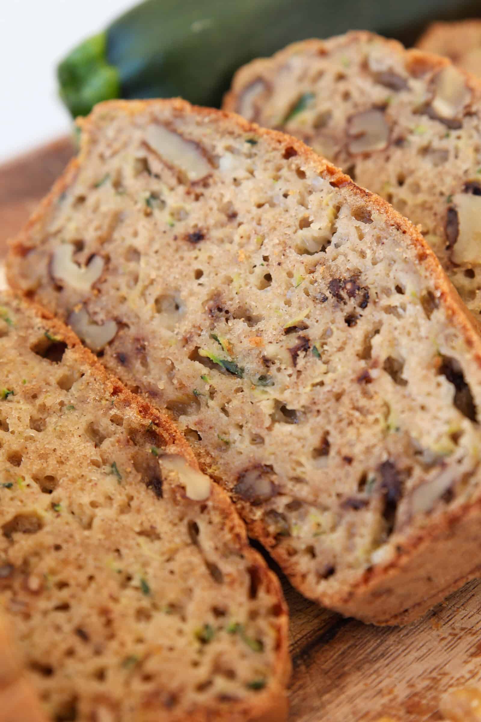 Close-up of sliced cottage cheese zucchini bread with visible pieces of walnuts and zucchini on a wooden surface, with a whole zucchini in the background.